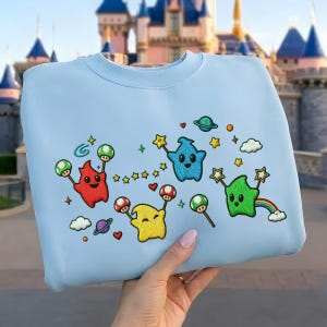 Puede incluir: Sudadera azul claro con personajes de dibujos animados bordados en rojo, azul, amarillo y verde. El dise&ntilde;o incluye estrellas, planetas, setas y un arco&iacute;ris. La sudadera la sostiene una persona.