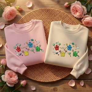 Puede incluir: Dos sudaderas de cuello redondo, una rosa y otra crema, se muestran sobre una estera tejida. Cada sudadera presenta un dise&ntilde;o bordado colorido de personajes de dibujos animados, estrellas y planetas. Rosas rosas y p&eacute;talos rodean las sudaderas sobre una superficie de madera.