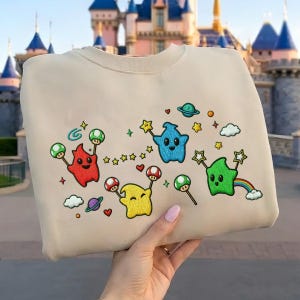 Puede incluir: Sudadera beige con personajes de dibujos animados bordados en rojo, amarillo, azul y verde. El dise&ntilde;o incluye estrellas, setas, planetas y un arco&iacute;ris. La sudadera es sostenida por una persona.