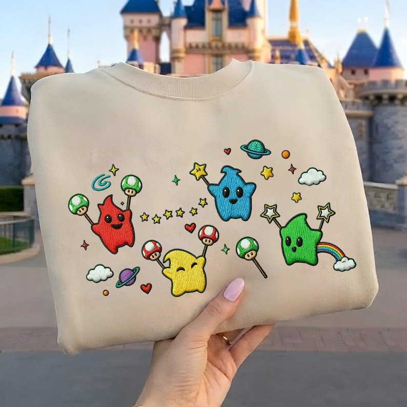 Puede incluir: Sudadera beige con personajes de dibujos animados bordados en rojo, amarillo, azul y verde. El dise&ntilde;o incluye estrellas, setas, planetas y un arco&iacute;ris. La sudadera es sostenida por una persona.
