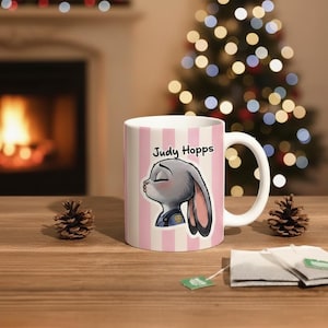 Peut inclure: Mug en c&eacute;ramique blanche &agrave; rayures verticales roses avec une image de dessin anim&eacute; de Judy Hopps de Zootopie. La tasse a une anse blanche et le texte "Judy Hopps" en noir. La tasse est sur une surface en bois avec des pommes de pin et des sachets de th&eacute;.