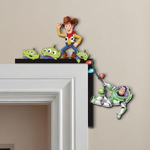 Puede incluir: Una pieza decorativa de esquina con personajes de la franquicia Toy Story. Woody, los extraterrestres y Buzz Lightyear están representados en colores vibrantes. La pieza está diseñada para encajar sobre un marco de puerta, agregando un toque lúdico.