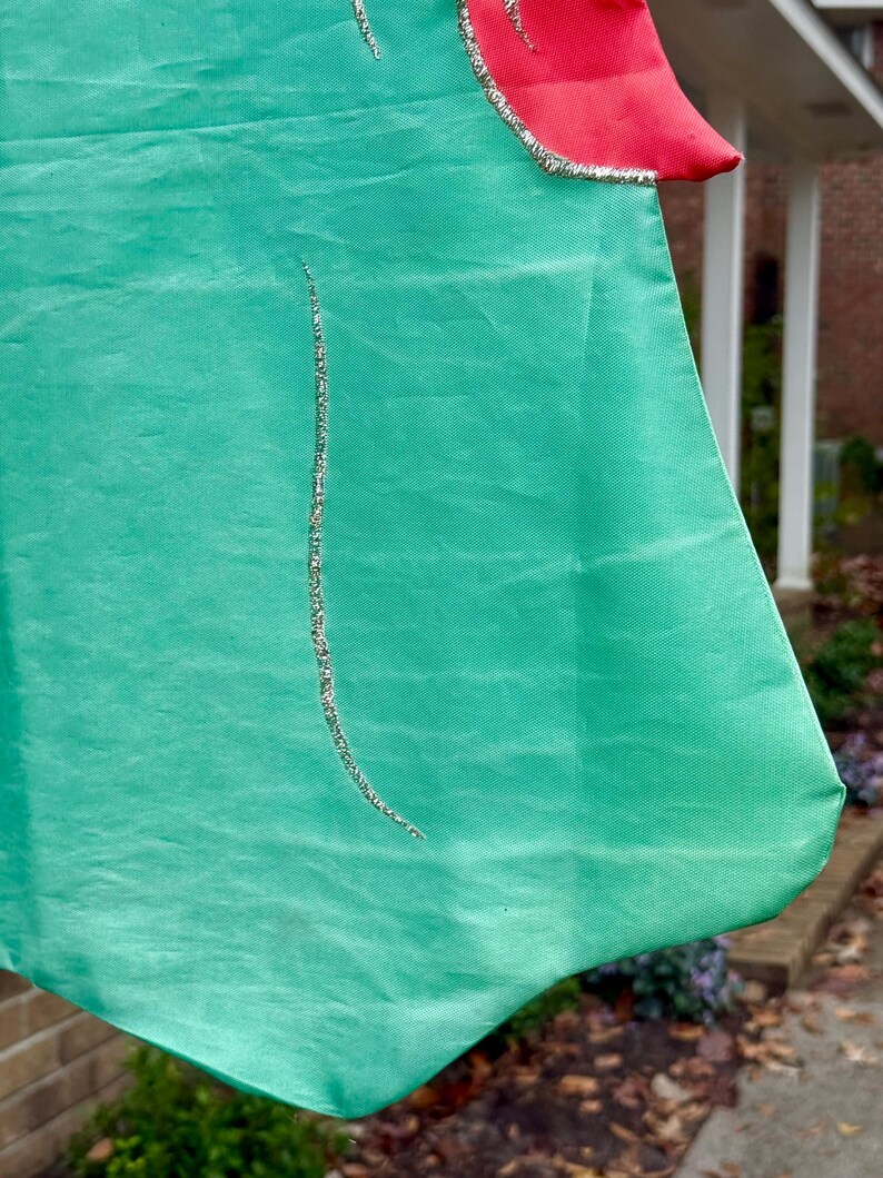 May include: A green fabric flag with a red accent at the top. Silver glitter trim outlines the red section and a vertical line down the green fabric. The flag is hanging outside.