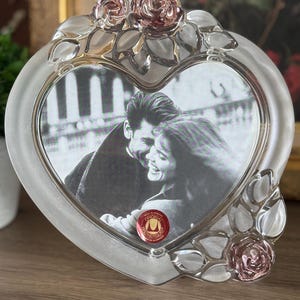 May include: A heart-shaped glass picture frame featuring frosted glass edges and clear glass accents. The frame is decorated with sculpted pink glass roses and leaves at the top and bottom right. A red circular sticker is visible on the glass.