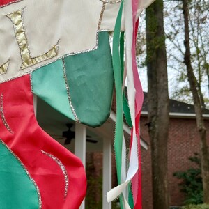 May include: A Christmas stocking with red, green, and white fabric, accented with gold trim and the letter "L". Colorful ribbons in green, white, and pink hang from the top. The stocking is outdoors.