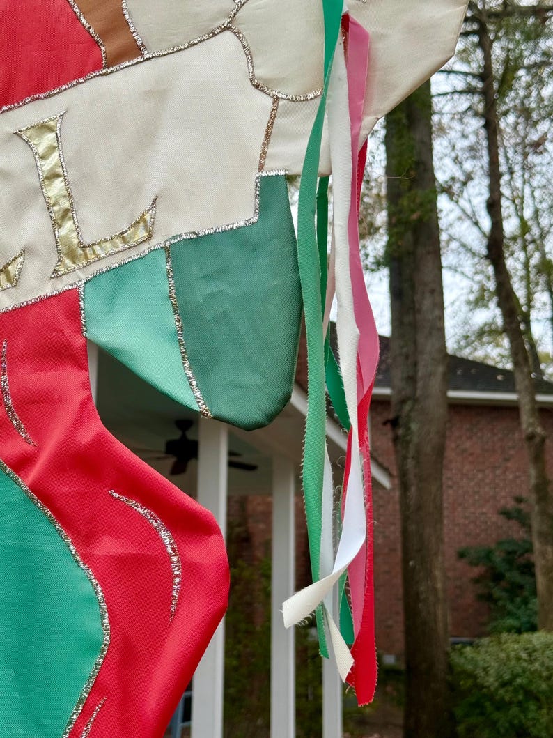May include: A Christmas stocking with red, green, and white fabric, accented with gold trim and the letter "L". Colorful ribbons in green, white, and pink hang from the top. The stocking is outdoors.