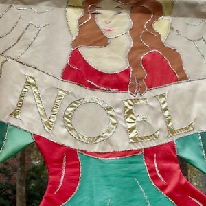 May include: A decorative Christmas banner featuring an angel with flowing auburn hair and a red dress. The banner reads "NOEL" in gold lettering. The angel's wings and the banner's edges are outlined with silver glitter.