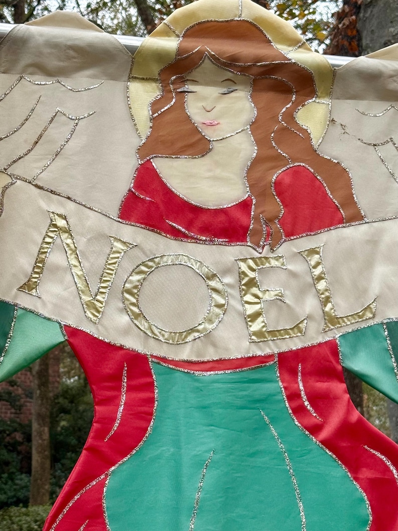 May include: A decorative Christmas banner featuring an angel with flowing auburn hair and a red dress. The banner reads "NOEL" in gold lettering. The angel's wings and the banner's edges are outlined with silver glitter.