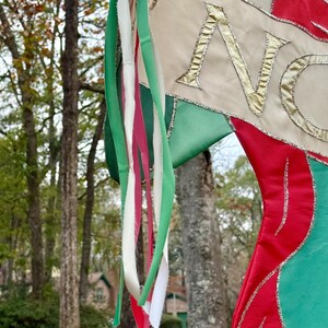 May include: A Christmas stocking with red, green, and cream-colored fabric. The word "NO" is embroidered in gold thread. Green, white, and red ribbons hang from the top, with a blurred outdoor background.