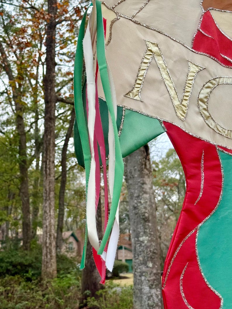 May include: A Christmas stocking with red, green, and cream-colored fabric. The word "NO" is embroidered in gold thread. Green, white, and red ribbons hang from the top, with a blurred outdoor background.