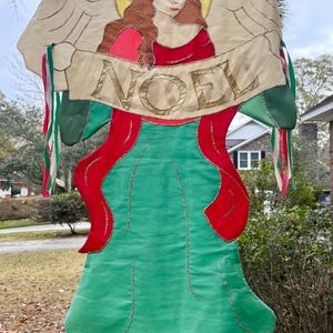 May include: A decorative Christmas banner featuring an angel illustration with the word "NOEL" in gold lettering. The banner has a green body with red accents and gold trim, and is adorned with colorful ribbons.