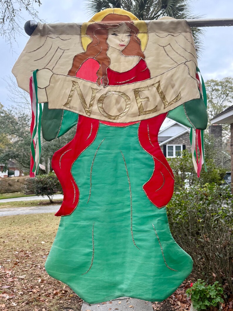 May include: A decorative Christmas banner featuring an angel illustration with the word "NOEL" in gold lettering. The banner has a green body with red accents and gold trim, and is adorned with colorful ribbons.