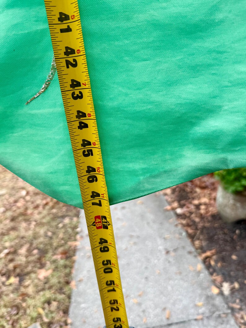 May include: A bright green fabric is measured with a yellow tape measure. The tape measure shows measurements from 41 to 53 inches. A small, gold-colored embellishment is visible on the fabric.