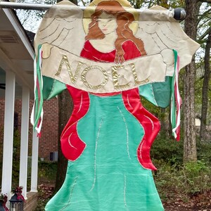 May include: A decorative Christmas banner featuring an angel design. The angel has red hair and a red dress, with a green skirt. The banner reads "NOEL" in gold lettering. The banner is trimmed with green and red ribbons.