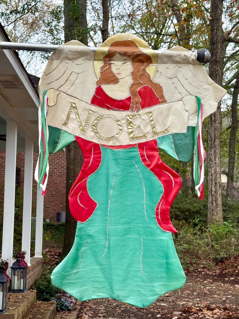 May include: A decorative Christmas banner featuring an angel design. The angel has red hair and a red dress, with a green skirt. The banner reads "NOEL" in gold lettering. The banner is trimmed with green and red ribbons.