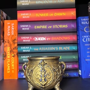 May include: A small, gold-colored cauldron with three clawed feet and two side handles. The front features a raised, circular emblem with an intricate, symmetrical design. Behind the cauldron are several stacked books by Sarah J. Maas.