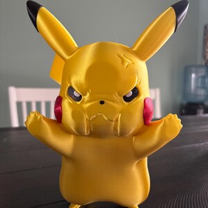 May include: A yellow Pikachu figurine with black-tipped ears, red cheek circles, and an angry expression. The arms are outstretched. The figurine is a 3D-printed item, likely a collectible or toy.