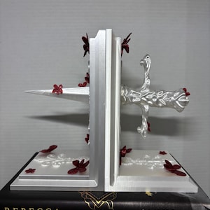 May include: A pair of white bookends with a decorative sword design and red floral accents. The bookends are sitting on top of a stack of books. The bookends are made of a 3D printed material.