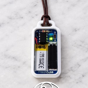 Può includere: Ciondolo rettangolare bianco con una finestra trasparente che rivela componenti interni, tra cui una batteria e un circuito stampato. Il ciondolo è sospeso a un cordino di cuoio marrone. La parola "Freehouse" è stampata.
