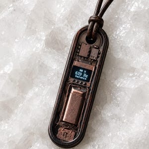 May include: A rectangular pendant necklace with a dark brown leather cord. The pendant has a copper-colored metal frame with internal electronic components and a small screen displaying "80 X 639 Hz balance".