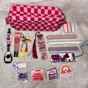 May include: A collection of reading accessories and stationery items, including a pink and white checkered pencil case, pens, bookmarks, stickers, and a bottle opener. The stickers have text that reads "Mood Reader", "main character energy", and "Romance Reader".