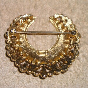 Puede incluir: Broche vintage dorado con dise&ntilde;o de semic&iacute;rculo. El broche presenta un patr&oacute;n calado con cierre de alfiler en la parte posterior. La parte posterior del broche tiene una marca del fabricante. El broche es probablemente de metal.