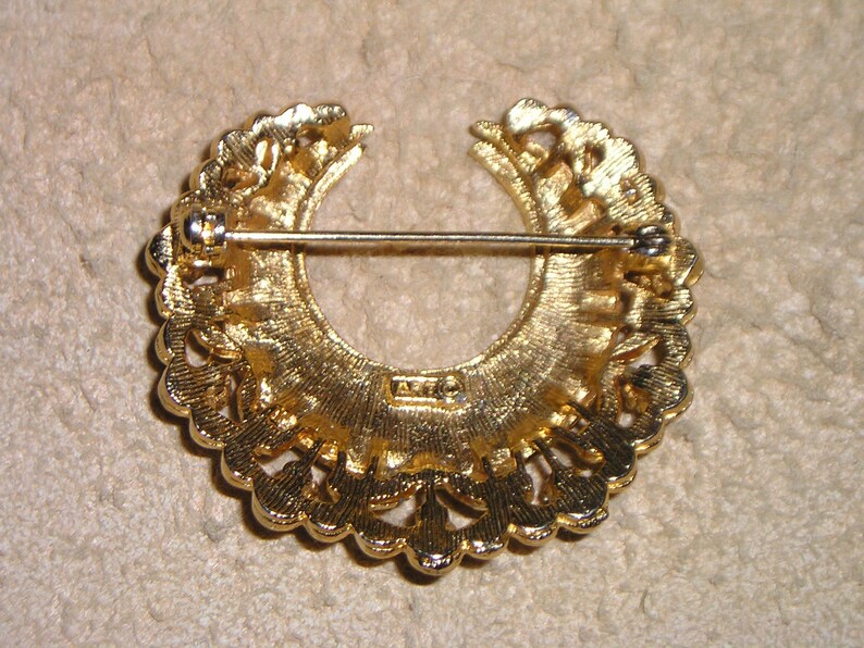 Puede incluir: Broche vintage dorado con dise&ntilde;o de semic&iacute;rculo. El broche presenta un patr&oacute;n calado con cierre de alfiler en la parte posterior. La parte posterior del broche tiene una marca del fabricante. El broche es probablemente de metal.