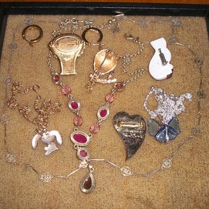 May include: A collection of vintage jewelry pieces, including necklaces, earrings, and brooches, displayed on a brown surface. The jewelry features a variety of colors, including gold, silver, and pink.