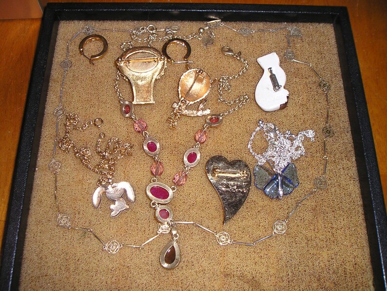 May include: A collection of vintage jewelry pieces, including necklaces, earrings, and brooches, displayed on a brown surface. The jewelry features a variety of colors, including gold, silver, and pink.