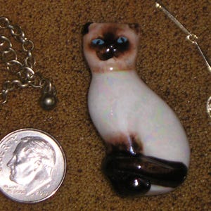 May include: A white ceramic Siamese cat figurine with blue eyes and brown markings. The figurine is sitting on a brown surface.
