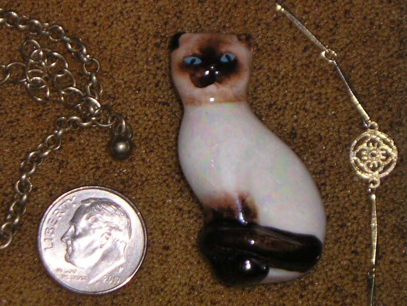 May include: A white ceramic Siamese cat figurine with blue eyes and brown markings. The figurine is sitting on a brown surface.