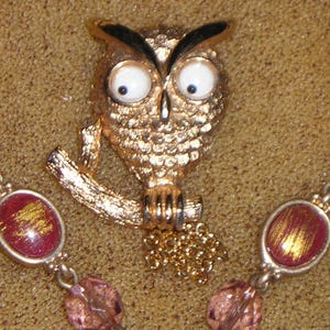May include: A gold owl brooch with large white eyes and black pupils. The owl is perched on a branch with a gold chain hanging from its feet.