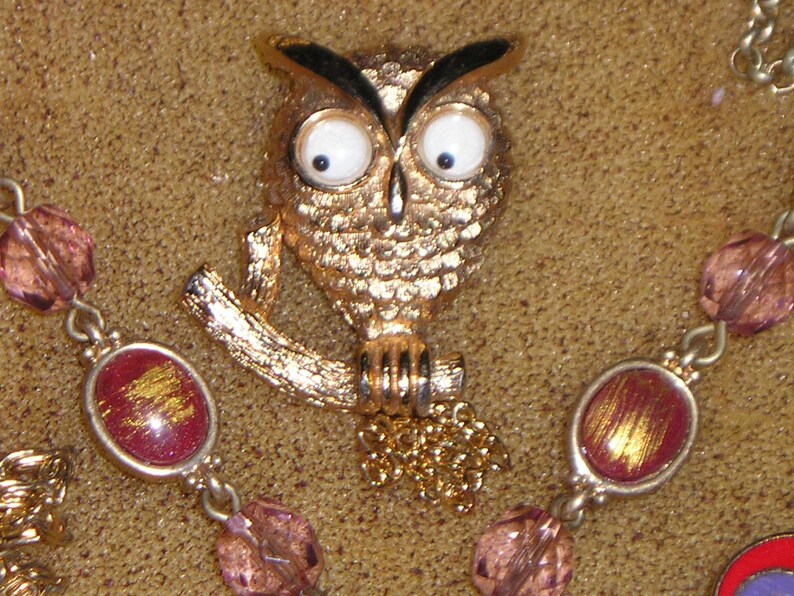 May include: A gold owl brooch with large white eyes and black pupils. The owl is perched on a branch with a gold chain hanging from its feet.