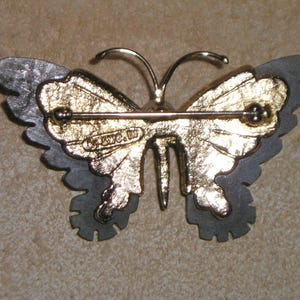 May include: A vintage butterfly brooch with a gold-tone centre and grey-tone wings. The brooch has a pin closure on the back and the text "&copy; S.W.C. W" is visible. The butterfly's wings have a layered design.