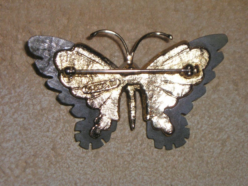 May include: A vintage butterfly brooch with a gold-tone centre and grey-tone wings. The brooch has a pin closure on the back and the text "&copy; S.W.C. W" is visible. The butterfly's wings have a layered design.