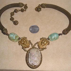 May include: An antique-style necklace with an Egyptian theme. The necklace features a woven gold-tone chain, turquoise scarab beetle beads, and gold-tone pendant with an oval central pendant. A U.S. quarter is next to the necklace for size comparison.