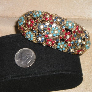 May include: A gold-tone bracelet with red, blue, and turquoise stones. The bracelet is on a black velvet cushion.