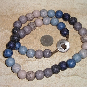 May include: A necklace composed of round, speckled beads in shades of blue, gray, and purple. The beads are strung together, forming a circular shape. A silver-colored button with the text "ATHENS DRO" is attached to the necklace.