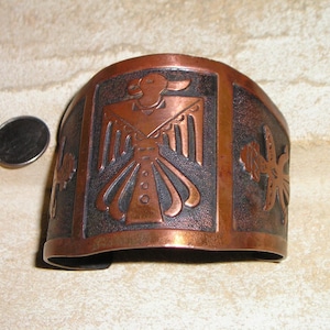 May include: A wide copper cuff bracelet featuring a central embossed thunderbird motif and side symbols. The metal has a dark, oxidized finish in the recessed areas and a polished copper tone on the raised surfaces, shown next to a US coin.