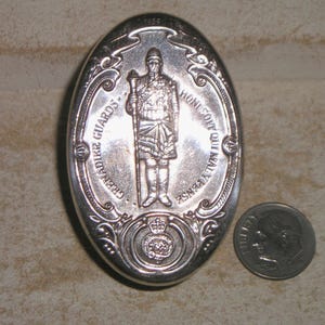 May include: An oval, silver-colored Grenadier Guards emblem with a detailed relief of a soldier holding a staff. The emblem is next to a dime for size comparison. The text "GRENADIER GUARDS" and "HONI SOIT QUI MAL Y PENSE" are visible.