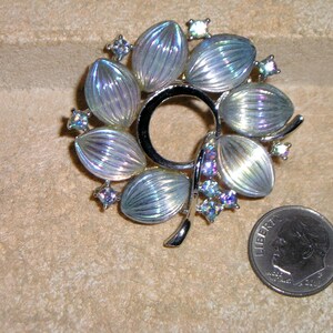 May include: A silver-toned floral brooch with iridescent, petal-shaped accents. The brooch features a central ring design and is adorned with small, sparkling crystals. A U.S. dime is included for size reference.