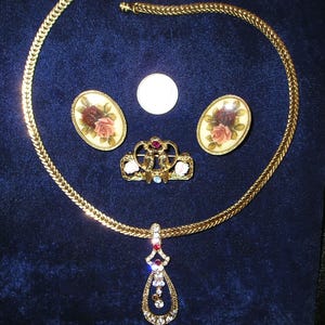 May include: A gold-toned jewelry set including a necklace with a teardrop pendant, a pair of oval earrings with rose designs, and a small brooch. The necklace has a woven chain. The pendant is adorned with clear and red gemstones.