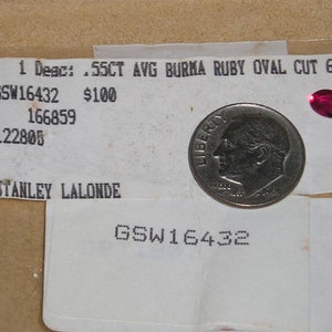 May include: A close-up of a small, oval-shaped, ruby gemstone, measuring 6mm by 4mm. The gemstone is a vibrant red color and is set against a white background. The text "55CT AVG BURMA RUBY OVAL CUT 6X4MM" is visible above the gemstone.