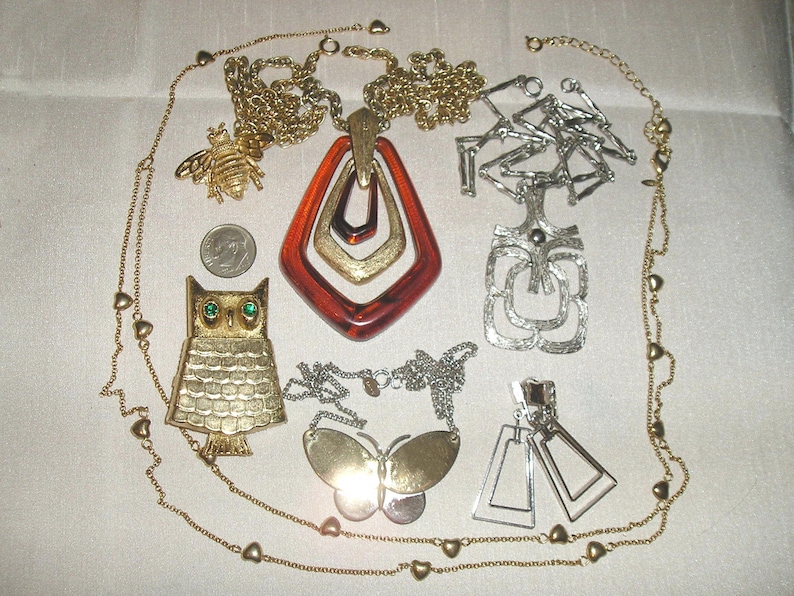 May include: A collection of vintage gold and silver tone necklaces with various pendants, including an owl, a bee, a geometric design, and a large amber-coloured pendant. The necklaces are on a white fabric background.