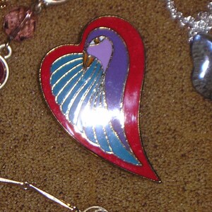 May include: A heart-shaped pin with a blue and purple bird design. The bird has a red beak and is surrounded by a red border.