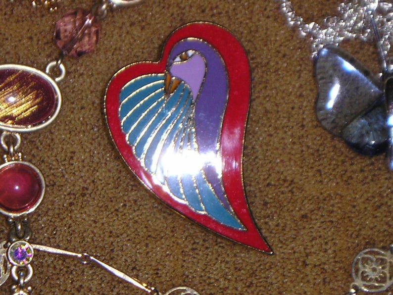 May include: A heart-shaped pin with a blue and purple bird design. The bird has a red beak and is surrounded by a red border.