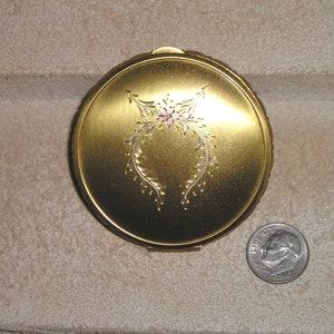 May include: A gold-toned compact with a floral design and a pink rhinestone in the center. The compact is about the size of a US dime.