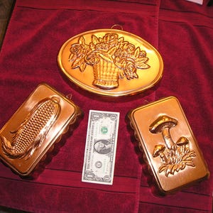 May include: Three copper molds: an oval floral basket, a rectangular corn, and a rectangular mushroom design. The molds are displayed on a red towel with a one-dollar bill for size reference. The molds are vintage and decorative.