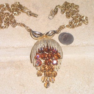 Puede incluir: Un collar de tono dorado con un gran colgante que presenta un grupo de piedras de imitación de color ámbar. El colgante tiene un diseño de hoja y una cadena de piedras de imitación más pequeñas que cuelgan de la parte inferior.