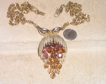 May include: A gold tone necklace with a large pendant featuring a cluster of amber-coloured rhinestones. The pendant has a leaf-like design and a chain of smaller rhinestones hanging from the bottom.